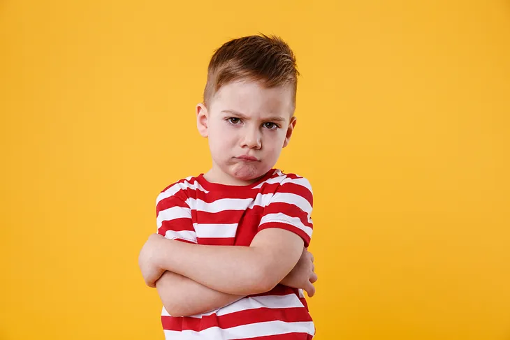 A grumpy-looking young boy in a red and white T-shirt against a yellow background. He has his arms folded across his upper body and looks quite cross.
