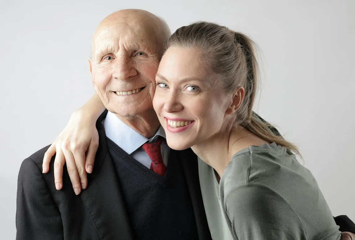 An attractive young woman has her arm around the shoulders of a well-dressed elderly man and both are smiling.
