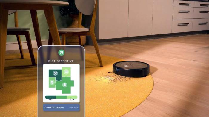 Marketing photo of the Roomba Combo J9+ robot vacuum. Overlay of the phone app in the foreground, showing its Dirt Detective map. The photo shows the robot in a kitchen / dining area on a yellow rug with dirt on it next to a dining set.