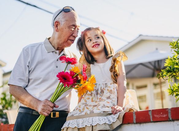 An older gentleman holds a bouquet of flowers in his right hand, while his left hand circles the waist of a young girl next to him who is presumably his granddaughter.
