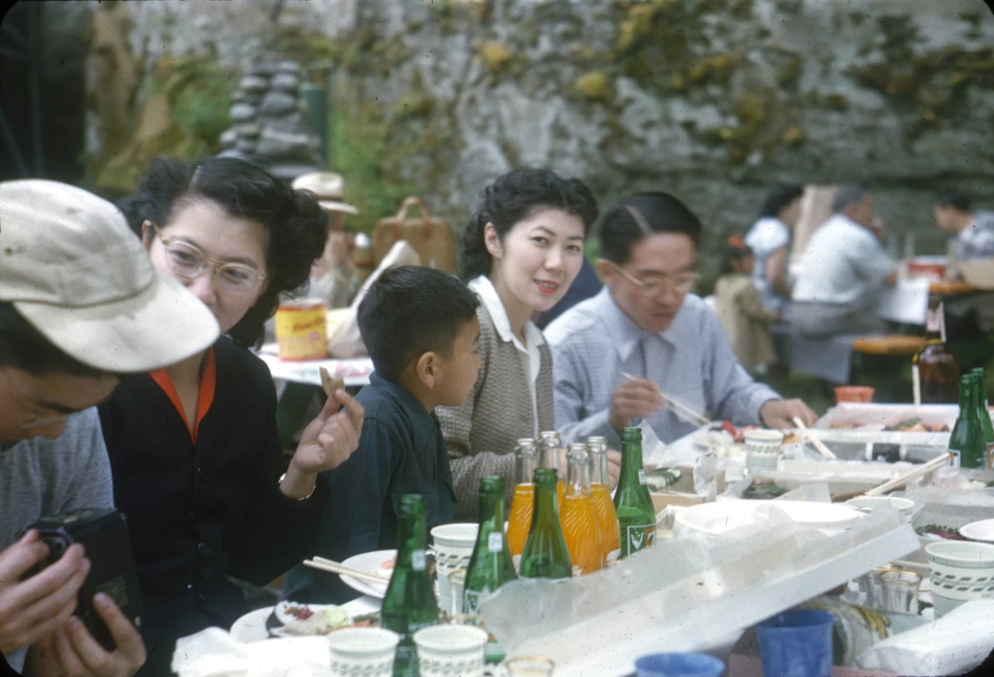 A group of people enjoying an outdoor gathering around a table filled with food and drinks, with one woman smiling towards the camera while the others are engaged in conversation.
