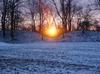 A landscape of snow covered grass, leafless trees over two symmetric mounds with a drop in the middle framing the sunrise.
