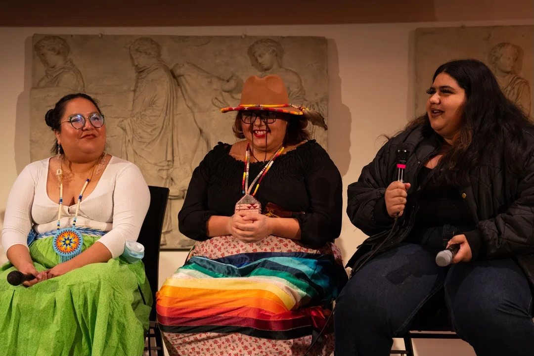 Three indigenous women engaged in a lively discussion at a panel event, set against a backdrop featuring classical relief artwork.
