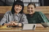 Two women smiling and sitting at a wooden table take a selfie with the Pixel Fold in Tabletop Camera mode.