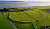 An aerial view of a large embossed circle in mowed green grass, with seawater in the distance.