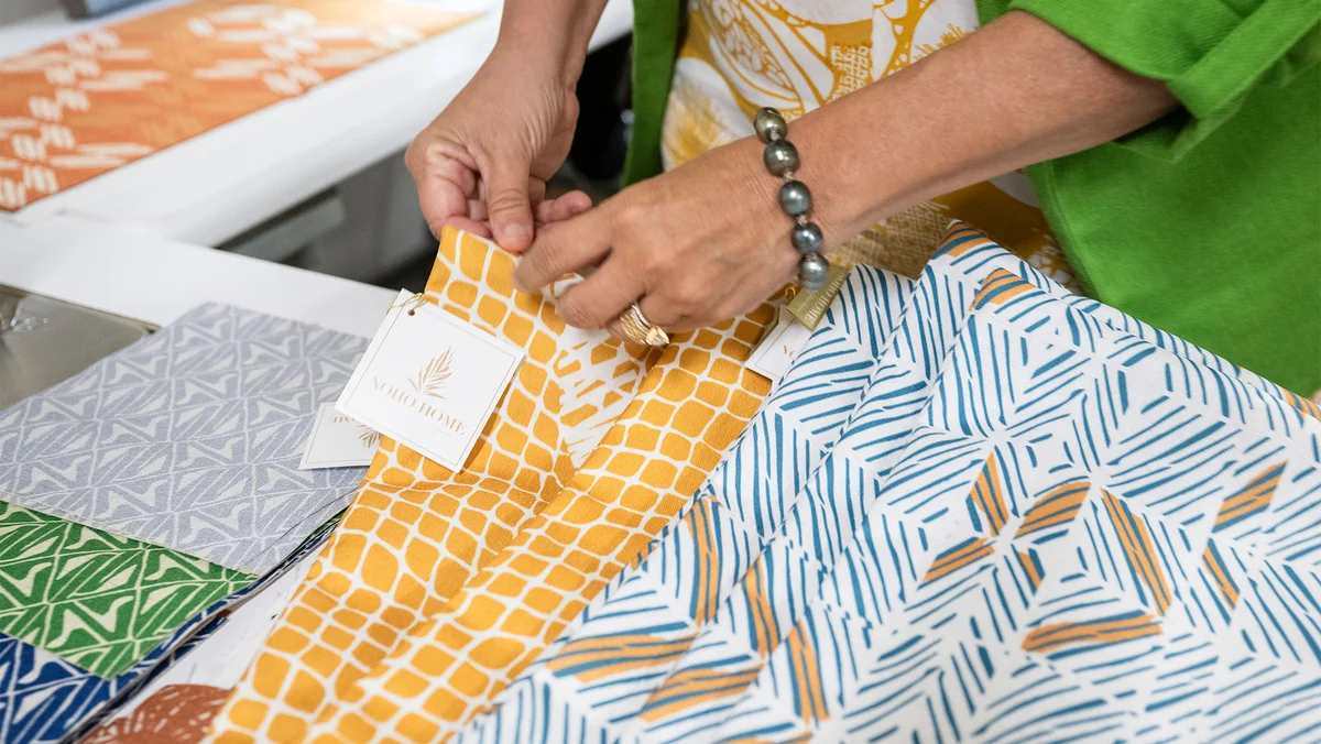 A close-up photo of hands working with various pieces of colorful fabrics. A tag on one piece of fabric reads "Noho Home."