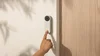 A hand comes into frame using the index finger to press a highlights button on a Nest Doorbell. The Doorbell is attached to the outside of a home, sitting next to a wooden door.