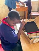 A young boy sits facing away from a desk as he opens up his new Chromebook with an excited grin