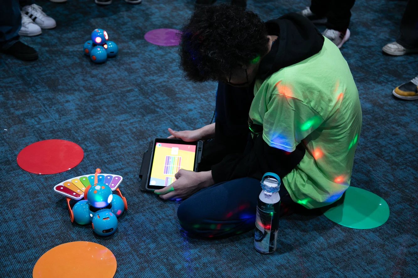 A male student kneeling on the floor uses a tablet device to write code for a robot that plays music.