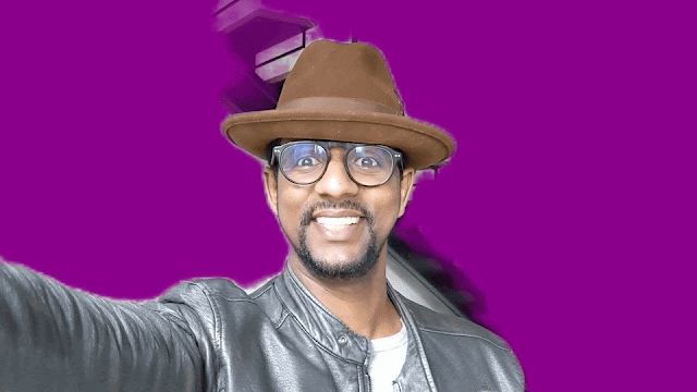 Animated gif of a man wearing glasses and a hat looking into a camera and waving and smiling. There's a green screen-like effect in the background, where everything behind him is purple but it appears to be concealing his actual settings. At first, the purple is only semi-successful at this; then you see the screen change and the purple more accurately surround the man and conceal his background.