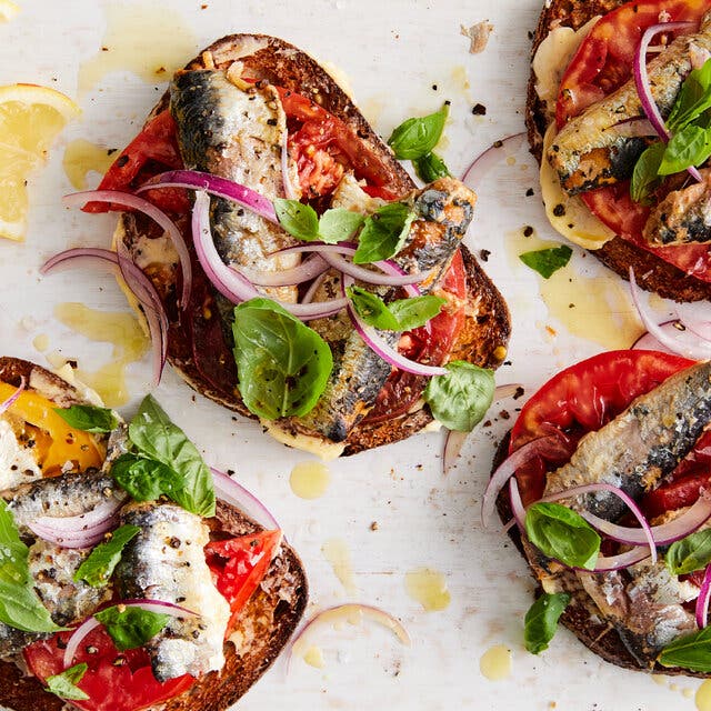 Four pieces of toast are piled high with tomato, sardines and red onion. A few lemon wedges sit off to the side.